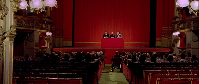 A paranormal conference, approached by Dario Argento's roving camera through red curtains.